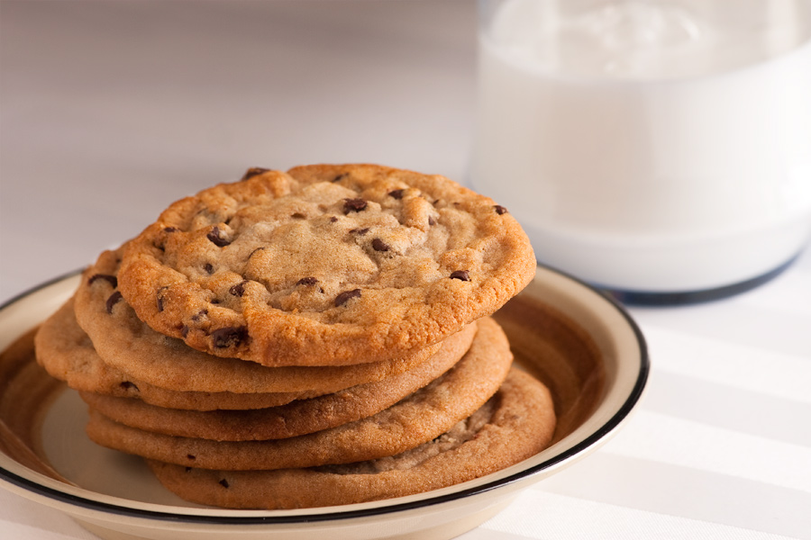 This Brian Charles Steel food photograph is of a stack of chocolate chip cookies and a glass of milk.  The cookies are in the front left portion of the frame on a light brown ceramic plate.  They are stacked at a slight angle extending out towards the glass of milk.  The glass of milk is in the back right portion of the frame.  Milk comes across as blue on camera, so the milk glass is actually filled with Elmer’s glue. 