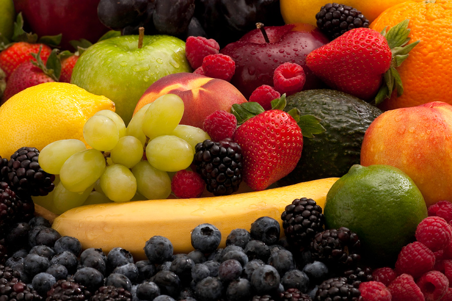 This is a Brian Charles Steel photograph of fruit.  Fruit fills the frame entirely.  The front is composed of blackberries, blueberries, and raspberries.  Behind the blueberries is a banana at a slight angle.  In front of the banana on the right side is a lime.  Behind the lime is a peach.  Behind the banana on the left are grapes and a strawberry.  Behind the grapes are a lemon and a peach.  Behind the peach are a green apple, a red apple and an orange.  The fruit has small drops of condensation and all of the fruit is in focus. 