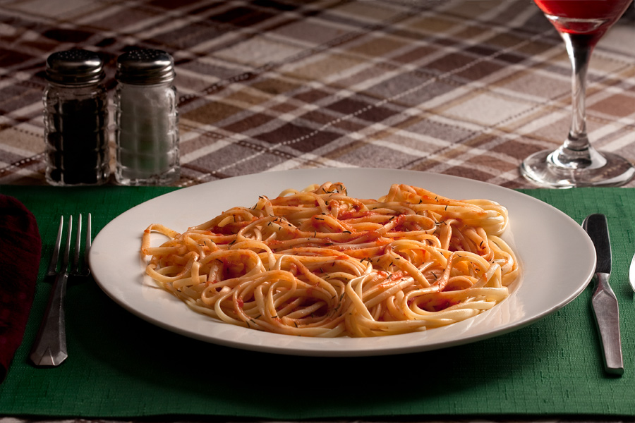 This is a Brian Charles Steel photograph of a plate of spaghetti on a brownish plaid tablecloth.  The white plate of spaghetti is sitting on a green placemat that is on the tablecloth.  The pasta has red sauce on it, and the noodles are neatly coiled, so that there are no visible loose ends.  The image was shot at a downward angle from above.  To the left and behind the plate are salt and pepper shakers.  On the sides of the plate are a fork, spoon and knife.  Behind the plate on the right side is a glass of red wine.  The pasta is lit from the back right side. 