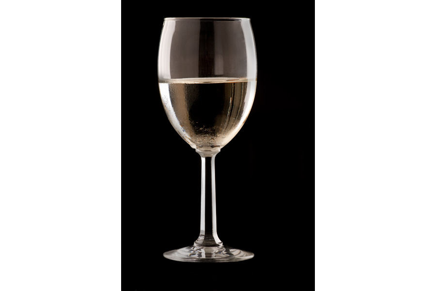This is a Brian Charles Steel photograph of a glass of white wine in a wine glass.  The image is a vertical shot, and the glass takes up most of the frame.  The glass is on a black background.  There are highlights on the far right and left sides of the glass.  There are beads of condensation on the glass. 