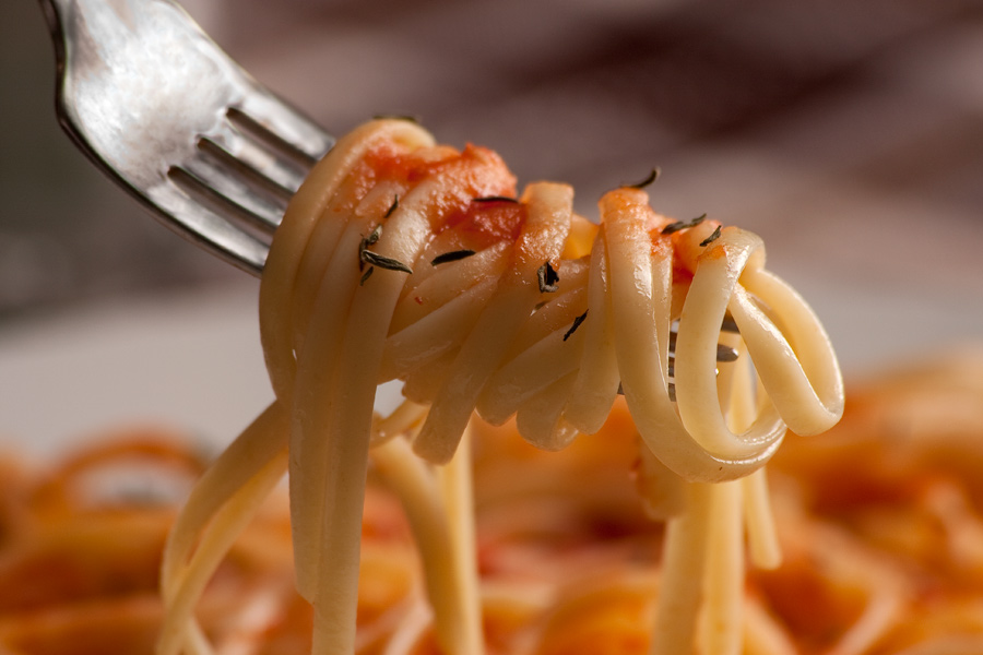 This is Brian Charles Steel close up shot of spaghetti wrapped around a fork.  The pasta is positioned toward the center of the frame.  The fork is positioned diagonally, and enters the frame from the top left corner.  Below and behind the subject is a plate of pasta that is out of focus.  Above that in the background is a brownish plaid tablecloth. 