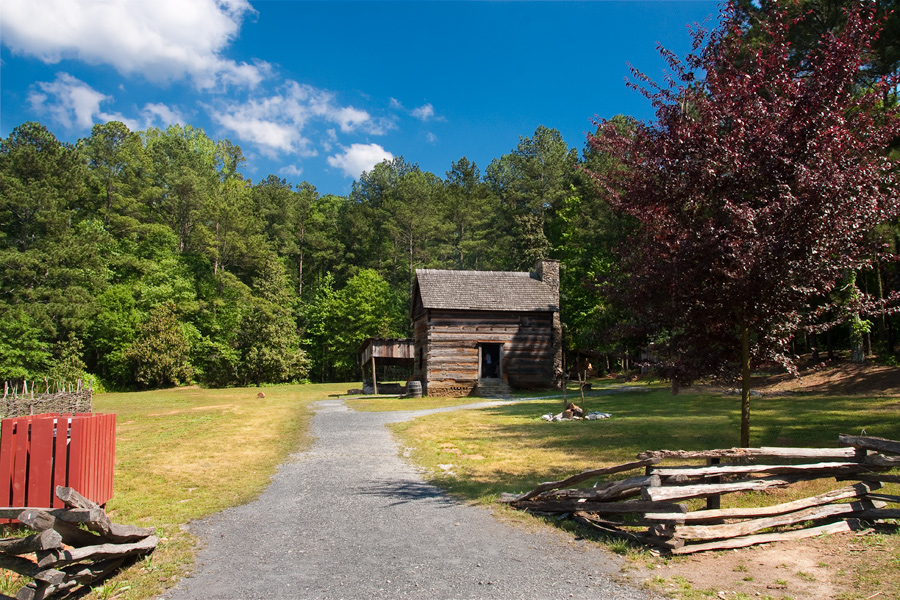 This is a Brian Charles Steel photograph of Fort Yargo.  There is a cabin in middle right portion of the frame towards the middle ground.  The cabin is made of logs and has one door.  It also has a brick chimney.   Behind the cabin there are tall green trees and bright blue sky.  Leading from the foreground to the cabin is a grey gravel road.  On each side of the gravel road there is a wooden fence in the foreground.  On the left side of the foreground is a red trashcan.  On the right side of the foreground there is tree with maroon leaves. 