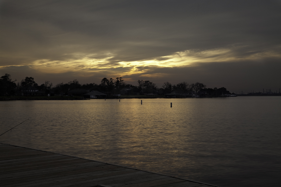 This Brian Charles Steel photo depicts Lake Prien in Lake Charles, Louisiana at sunset.  The bottom left portion of the frame shows the dock at a forty-degree angle.  The yellow-orange sunlight reflects off the water.  There are a series of buoys towards the middle of the frame.   There is stretch of land beyond the buoys; the land contains trees and houses.  The sky takes up the top half of the frame.  The sky is a mix of blue and red clouds.  In the middle of the sky is a streak of bright yellow orange light.  