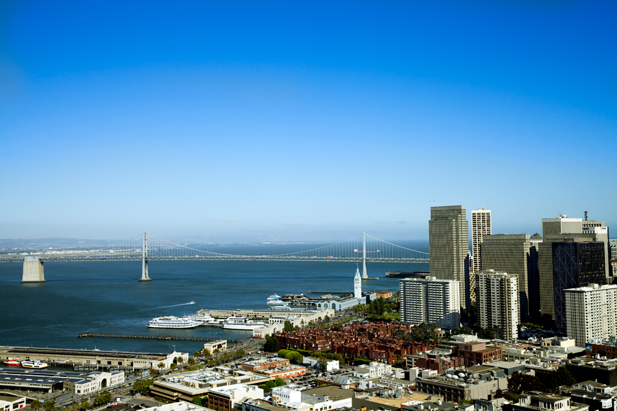 This Brian Charles Steel photo depicts the San Francisco Bay.  The photo is at an angle of that the water line makes a diagonal through the bottom third of the frame.  The diagonal starts in the lower left corner and works its way up to the top of the bottom third of the frame on the right.  There are white boats docked along the water’s edge.  On the right you can see the city skyscrapers.  To Oakland Bay bridge goes straight across just below the middle of the frame.  The sky takes up the top two thirds of the frame.  The sky is a very light blue on the bottom and a vibrant blue on the top. 