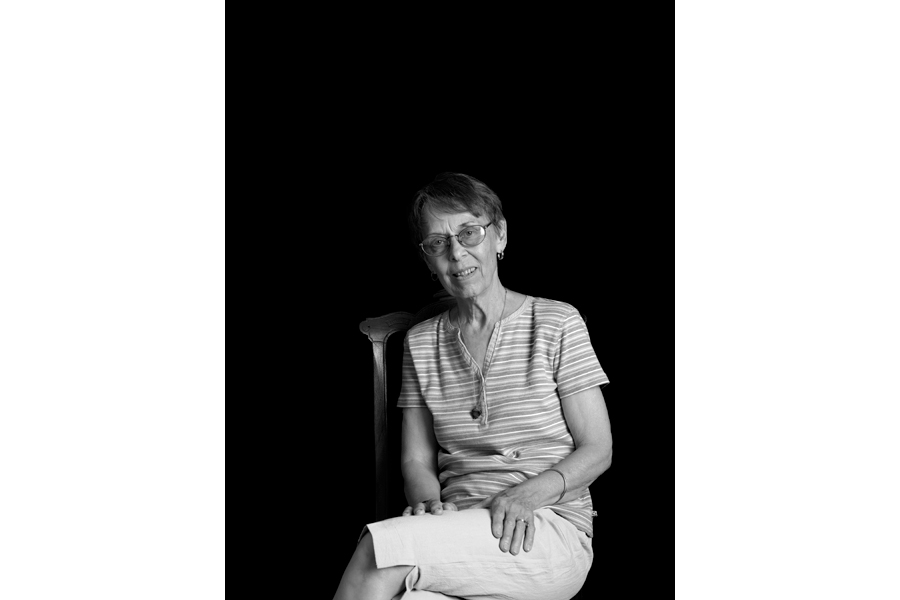 This is a Brian Charles Steel black and white photographic portrait of Sandy Brauer sitting in a chair.  She is in the lower middle portion of the frame.  She is shown from just below the knees and up.  She is wearing a short sleeve shirt with horizontal stripes and plain white shorts that go to the knees.  She has her legs crossed facing toward the left.  Her upper body is facing forward and she is looking into the camera.  Her hands are on her legs.  She has short brown hair and is wearing glasses.  The background is solid black. 