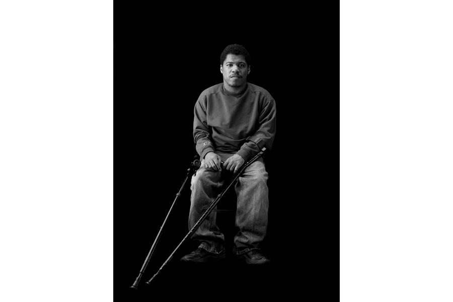 This is a Brian Charles Steel black and white photographic portrait of Charlie White.  Charlie is a thirty-year-old African American with cerebral palsy. His full body is in frame and takes up about one third of the image.  He is wearing a long sleeved sweater and blue jeans.  He is sitting in a chair towards the center of the frame.  His crutches are pointed diagonally towards the bottom left corner.  He is lit in a Rembrandt style with the main light source coming from the left.  He is looking directly into the camera. 