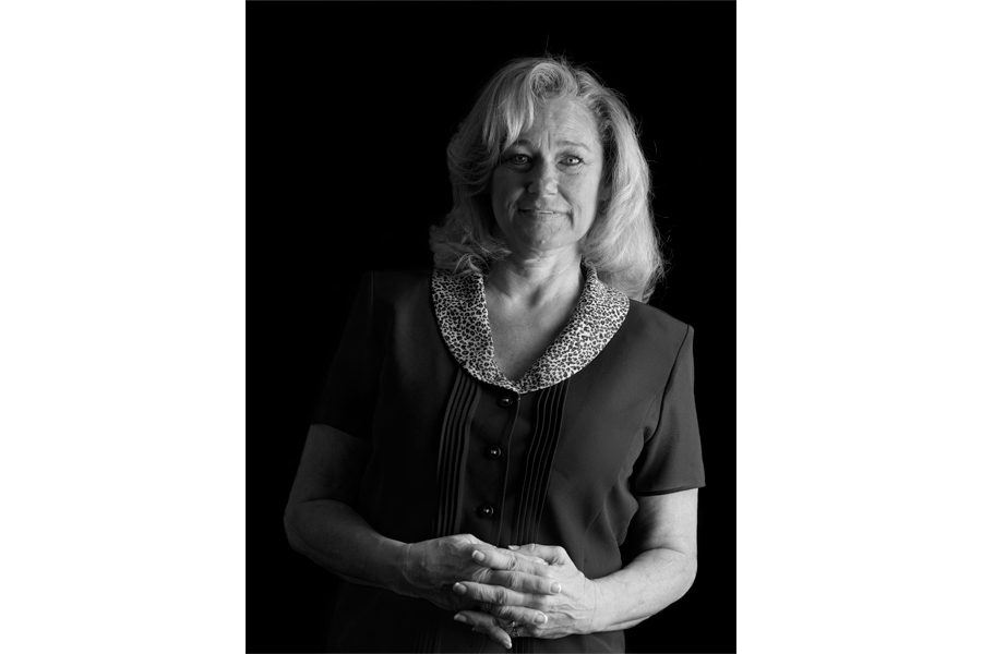 This is a Brian Charles Steel black and white photographic portrait of Allison Kerr.  She is positioned toward the center, and lit with a Rembrandt style.  The main light source is coming from the right.  She is shown from the waist up, and she looks directly into the camera.  Kerr has shoulder length blond straight blond hair.  She is holding her hands together over her stomach.  Kerr is wearing a short-sleeved button up shirt. The background is solid black. 