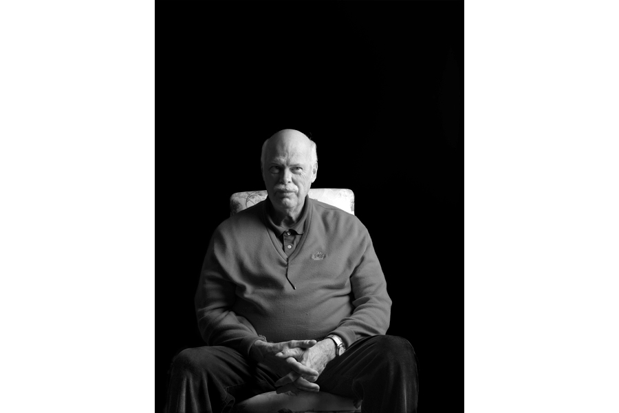 This is a Brian Charles Steel black and white photographic portrait of Hobo Bodkin.  He is seated in a chair and comprises the middle of the bottom half of the frame.  His hands are folded neatly in his lap.  He is wearing a red sweater and black corduroys.  Bodkin is lit with a Rembrandt style, and the light is coming from the right side.  His body is facing forward, and he is looking straight into the camera.  He is bald with a grey mustache. The background is solid black. 