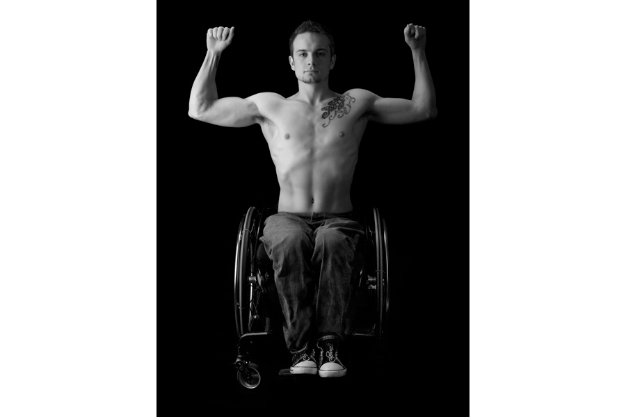 This is a Brian Charles Steel black and white photographic portrait of Richard Corbett.  He is positioned in the center of the frame.  He is flexing his arms up in the air in a bodybuilding pose.  He is seated in a wheel chair while wearing blue jeans and no shirt.  He is lit in a Rembrandt style with the main light coming from the left.  The light is soft. 