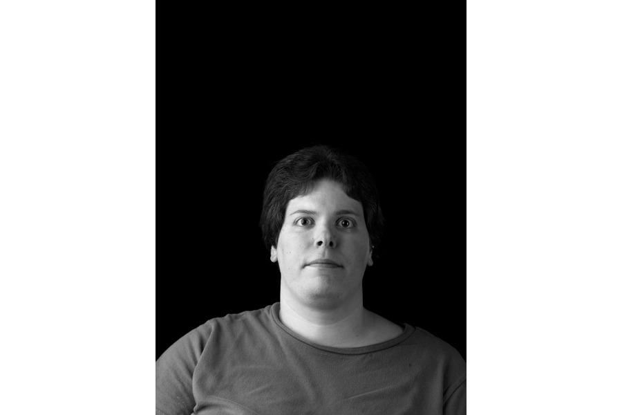 This is a Brian Charles Steel black and white photographic portrait of Tiffany Coleman.  She occupies the bottom half of the frame, and is shown from the chest up.  She has short dark hair, and is wearing a grey shirt.  Coleman is facing forward, and looking straight into the camera.  She occupies about one third of the frame. Coleman is lit with Rembrandt lighting with the main source coming from the left.  The background is solid black. 