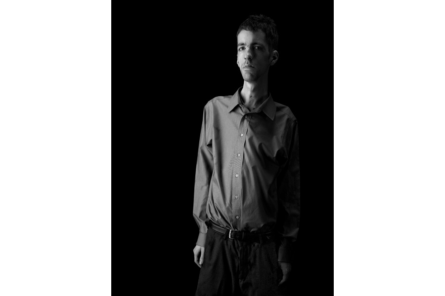 This is a black and white photographic portrait of photographer Brian Charles Steel.  Steel has congenital fiber type disproportion, which causes him to be thin and small framed.  Steel is positioned on the right side of the frame and he fills it from top to bottom on that side.  You see him from just above the knees and up.  He is wearing a long sleeved dress shirt with dress slacks.  His arms are at his side and he is looking straight into the camera.  He is lit in a Rembrandt style with the main light source coming from the left. 