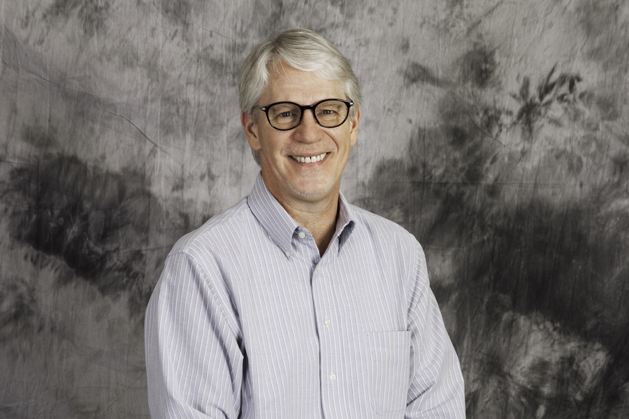 This Brian Charles Steel Photograph is a portrait of a man with grey hair in a blue and white striped shirt. He is seated in front of a grey background.  He is also wearing glasses. 
