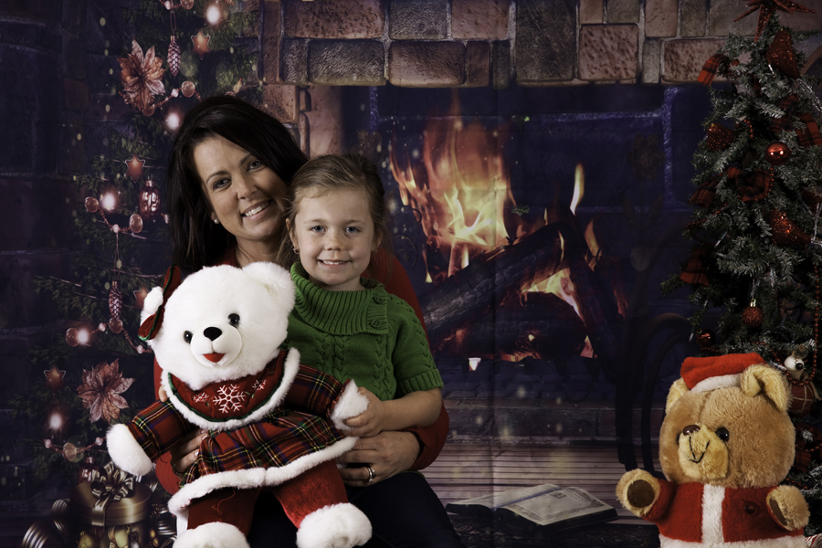 This Brian Charles Steel photograph is a Christmas portrait of a woman and her daughter.  Behind them is a brick fireplace with a roaring fire. On the left side of the background is a Christmas tree, and on the right is a teddy bear dressed like Santa.  The woman has long black hair, and the little girl has light brown hair.  The girl is holding a white teddy bear wearing a red and green plaid dress. 