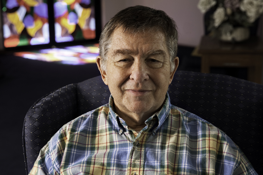 This Brian Charles Steel photo is a portrait of a retired priest.  He is seated in a chair. It is an upper-body shot.  He is wearing a plaid collared shirt.  Behind him to the left is a stained glass window.  