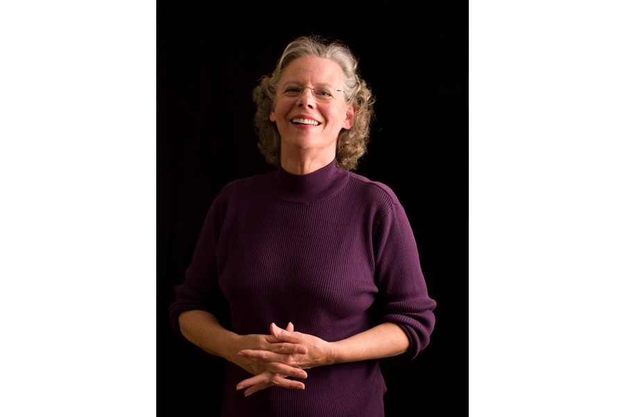 This is a Brian Charles Steel photographic portrait of Carolyn Caskey.  She is shown from the waist up, and she is holding her hands together over her stomach.  Her body is facing toward the left side of the frame and her head is turned forward looking into the camera.  She is lit with a Rembrandt style and the light source is coming from the right side.  Her hair is curly, and almost shoulder length.  She is wearing glasses.  The background is solid black. 