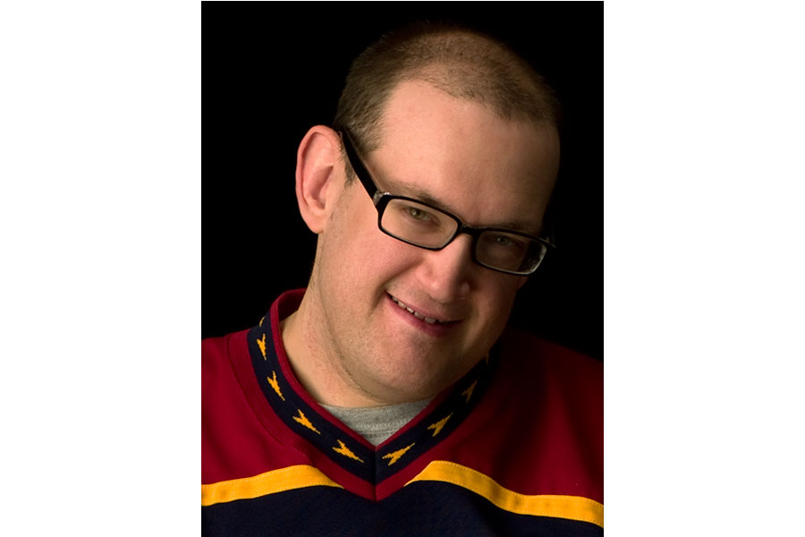 This is a headshot of Ben taken by Brian Charles Steel.  He is wearing glasses and a Thrashers jersey.  He is lit Rembrandt style with the main source coming from the left.  He looks directly into the camera.  His hairstyle is a buzz cut.  The background is solid black. 