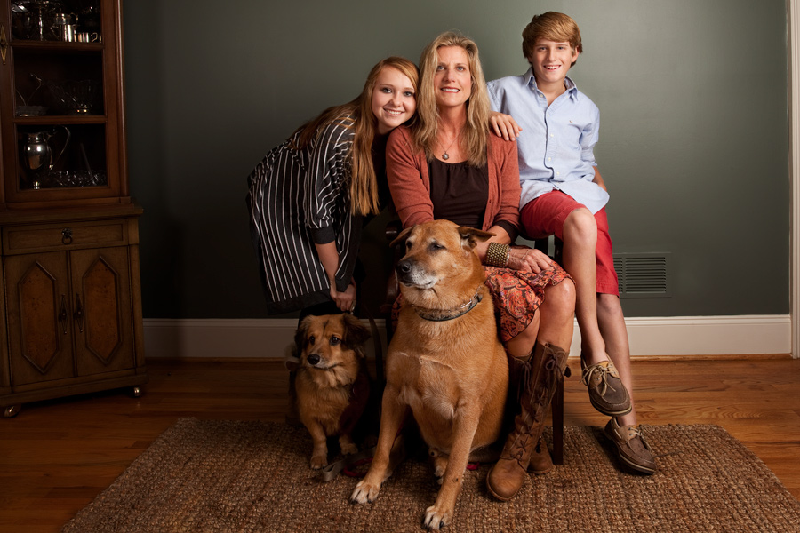 This is a Brian Charles Steel color photograph of a mother with her son, daughters, and two dogs.  The family is in the center of the frame.  Behind them is a dark green wall with white trim.  In the left back corner is a wooden china cabinet.  The floor is hard wood.  The mother is seated in a chair with her daughter to her left and son to her right. The dogs are in the floor in front of them.  The woman is thin with blond hair and is in her forties.  The son has short light brown hair and is around twelve.  The daughter has long brown hair and is about sixteen.  They are all huddled together and compose about half of the frame. 