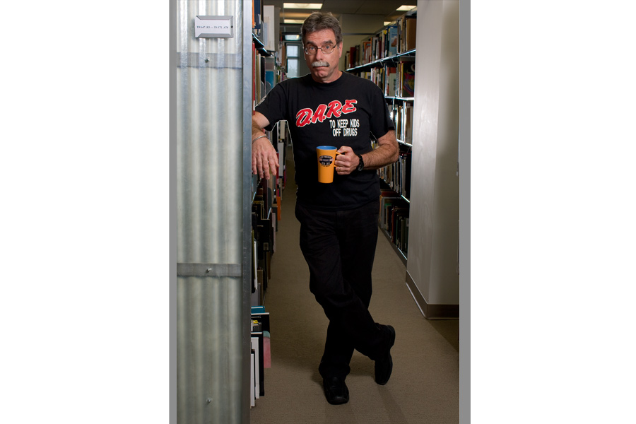 This is a Brian Charles Steel portrait of photographer Forest McMullin.  He is in a library between to book aisles.  He is wearing a black Dare shirt, and holding a coffee cup. His body is completely in frame, and he comprises about half of the frame.  He is positioned in the center. 