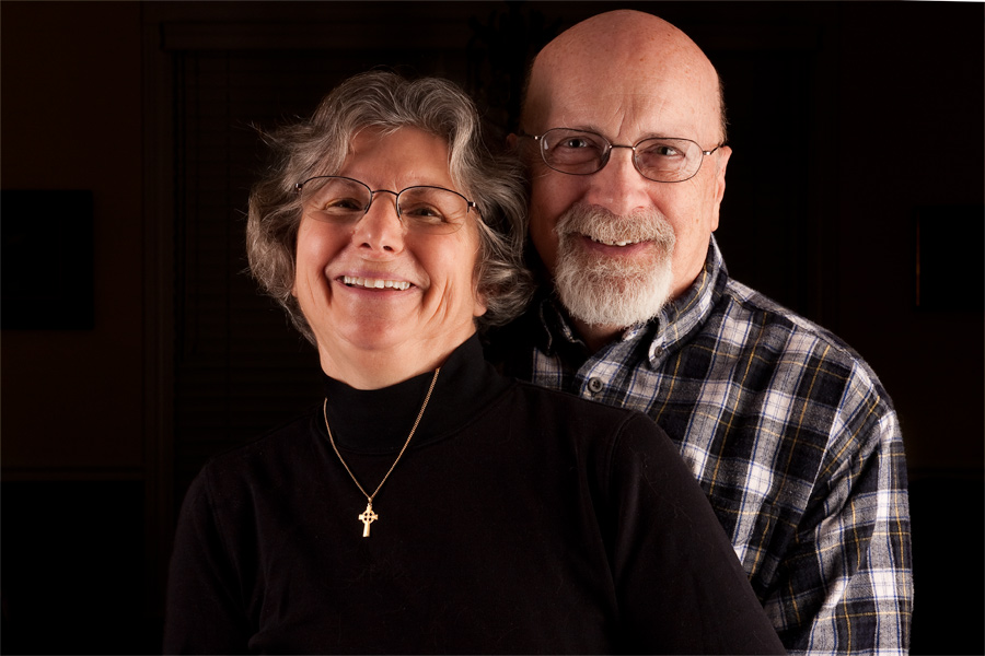 This is a Brian Charles Steel photographic portrait of John and Hazel.  They are toward the center of the frame.  John is wearing a plaid shirt and Hazel is dressed in black with a gold necklace. They are smiling.  They are shown from the chest up and comprise about three fourths of the frame.  They are lit from the front right side. 