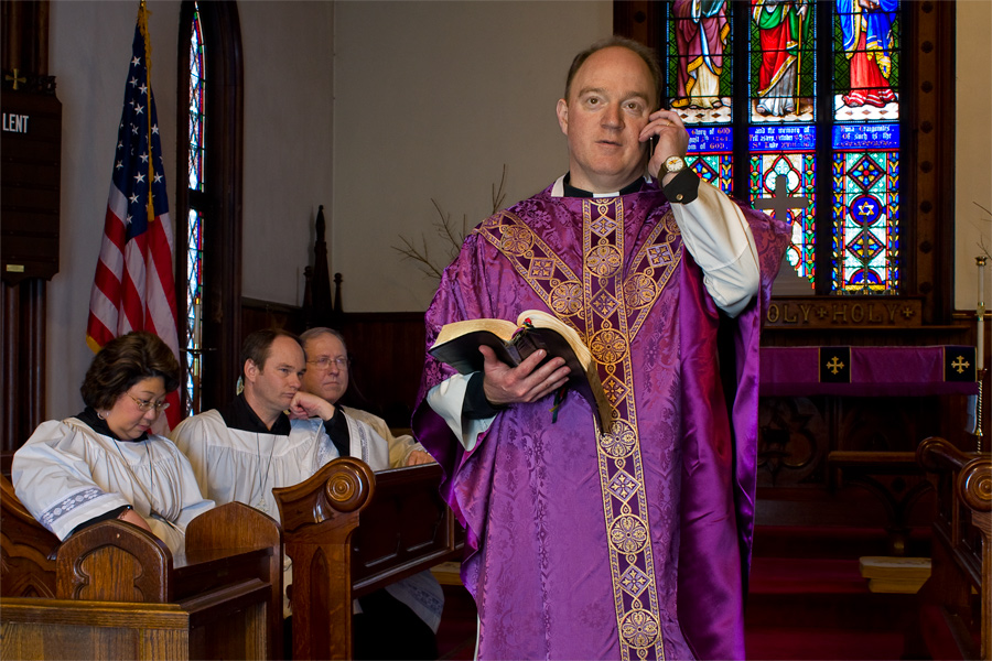 This is a Brian Charles Steel portrait of an Episcopal priest talking on his cell phone during a service.  He is in the center of the frame.  There is stained glass behind him to the right. To the left of him are choir members who appear to be frustrated him for being on the phone.  He holds is bible in his right hand and a phone in his left. 