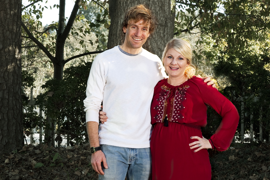 This Brian Charles Steel photo depicts a married couple celebrating their pregnancy with a baby shower.  They are standing in their yard with trees in the background. The husband is on the right and wearing blue jeans and a white shirt.  The wife is wearing a red dress, and they have their arms around each other as they look into the camera. Strobe flash causes them to pop out from the background. 