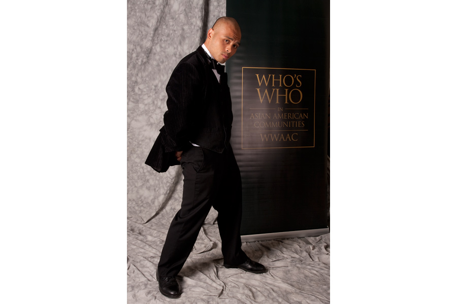 This Brian Charles Steel photo depicts a young Asian gentleman in front a grey back drop. He is wearing a tuxedo, and has his side turned to the camera.  Behind him is a banner for the Who’s Who in Asian American Communities.  