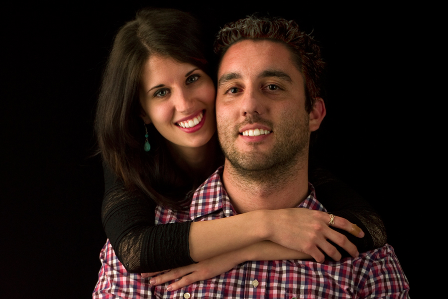 This is a Brian Charles Steel photograph of Kyle and Nikki.  Nikki is behind Kyle with her arms around him while hanging over his shoulders with her face beside his. They are both smiling, and looking into the camera.  She has long brown hair, and he has short curly hair.  They are towards the right side of the frame, and are seen from the chest up.  The background is solid black. 